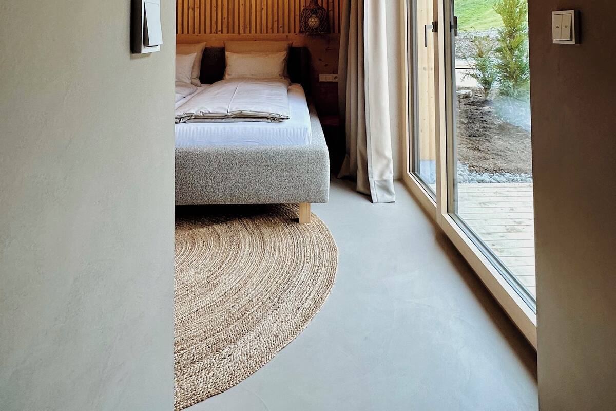 Une chambre moderne avec un grand lit, un mur lambrissé en bois, un luminaire au plafond et une vue sur un paysage verdoyant, visible à travers une porte-fenêtre coulissante.
