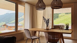Un comedor con mesa de madera, sillas de mimbre y vista a un paisaje verde a través de ventanas grandes.