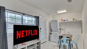 Une pièce dotée d’un grand téléviseur à écran plat affichant le logo « NETFLIX », d’une petite kitchenette équipée d’un four à micro-ondes, d’une table avec des chaises et d’une fenêtre avec des rideaux.
