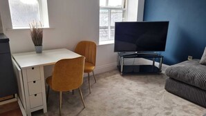 A room with a white desk, two yellow chairs, a black TV on a stand, a grey sofa, and a window with a view of buildings. - Cosy, Central Apartment Skipton (Skipton)