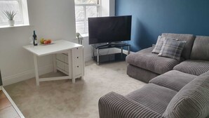 A living room with a white desk, a flat-screen TV, a grey sofa, and a blue accent wall. - Cosy, Central Apartment Skipton (Skipton)