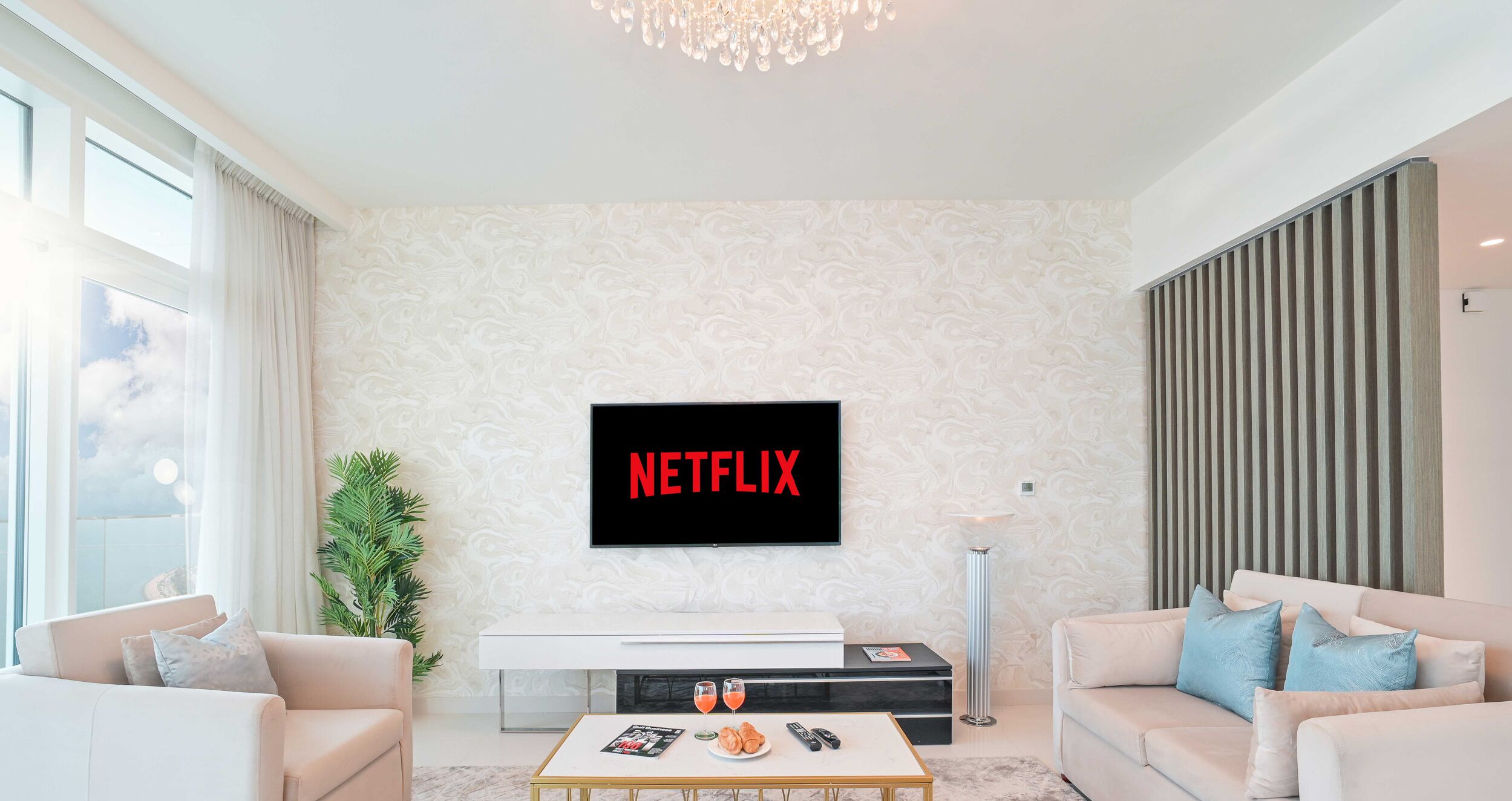 Ein modernes Wohnzimmer mit einem Flachbildfernseher, der "NETFLIX" anzeigt, einem weißen Sofa, einem Couchtisch mit Zeitschrift und Gläsern sowie einer hohen Stehlampe.