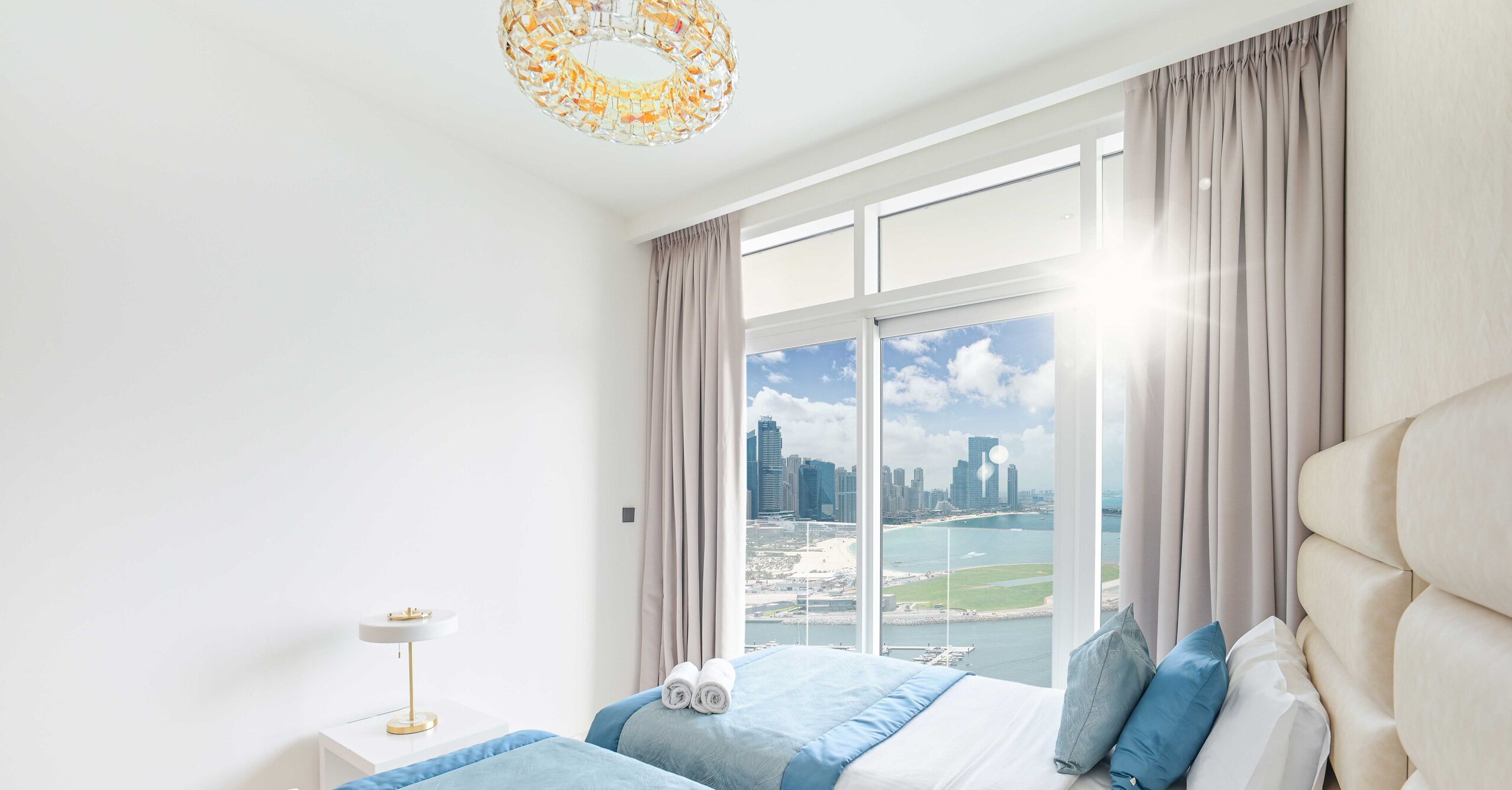 A modern hotel room with a large window offering a city view, a bed with blue bedding, and a white bedside table with a lamp.