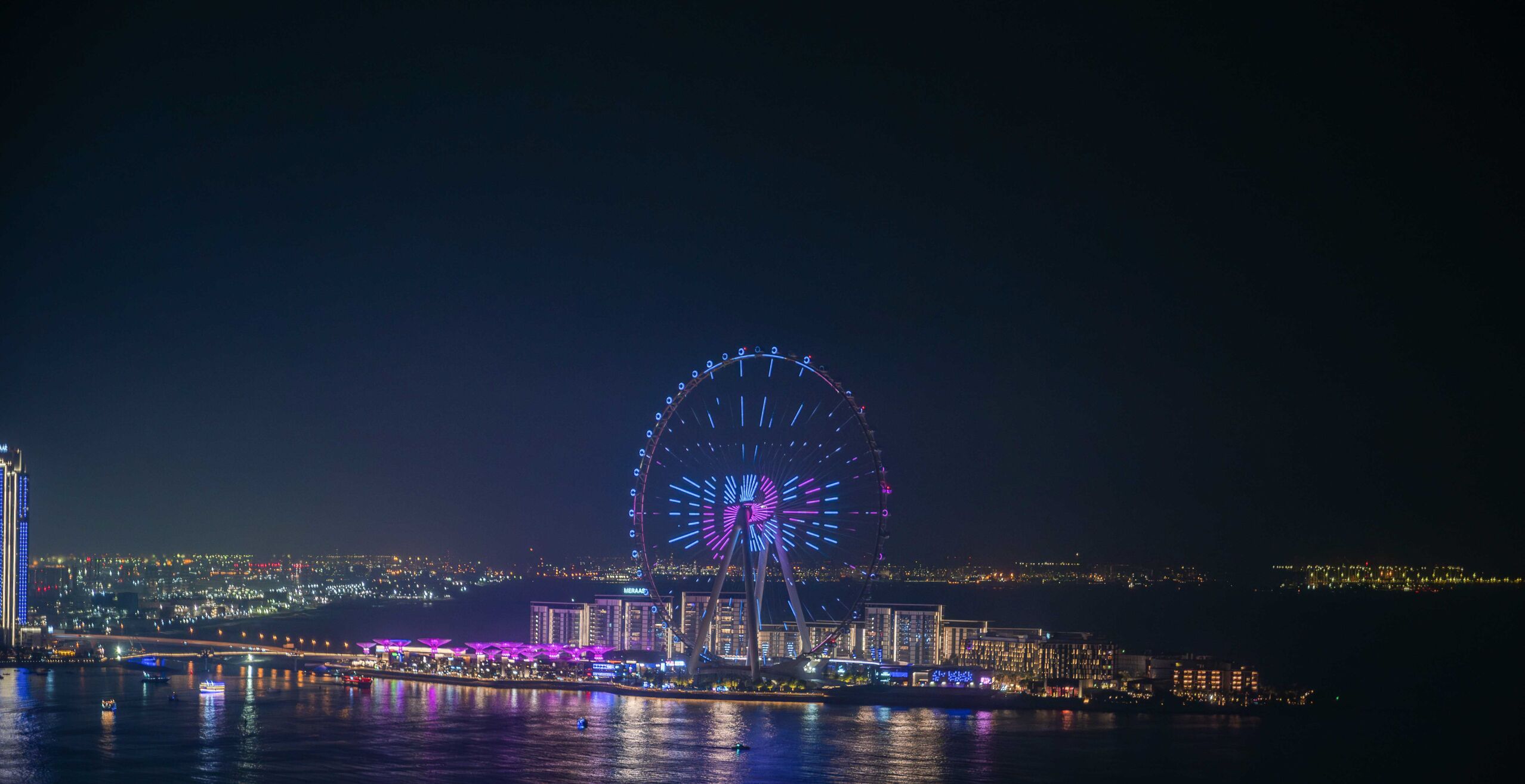 Una noria grande con luces de colores, un frente marítimo y edificios iluminados al fondo.