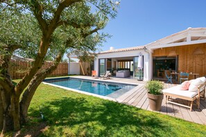 Un jardin avec une piscine, une terrasse en bois, du mobilier d’extérieur et un grand arbre.