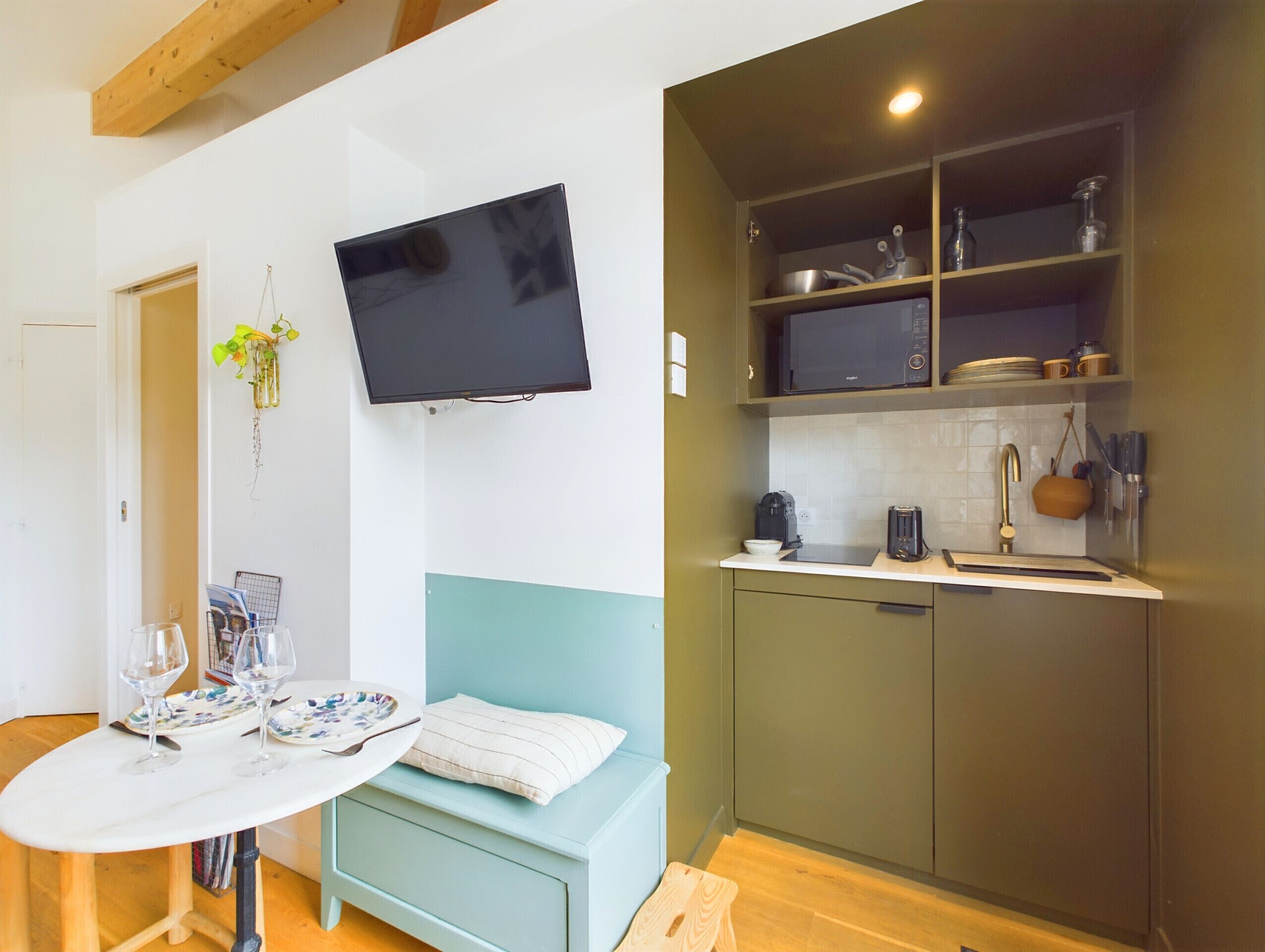 A compact kitchen with a microwave, sink, and cabinets. A flat-screen TV is mounted above a dining table with two wine glasses.