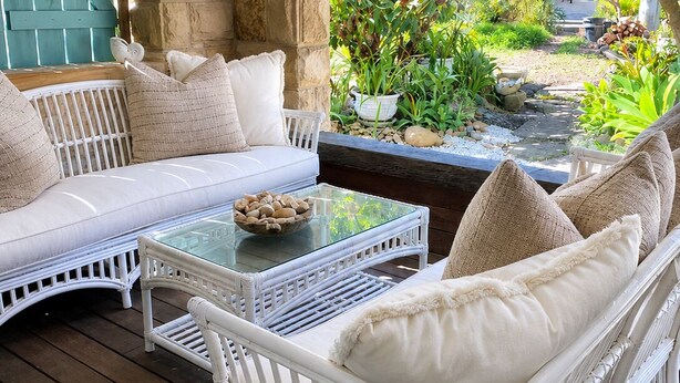 Un conjunto de sofá y sillón de mimbre blanco con cojines, una mesa de centro con superficie de vidrio y un fondo de pared de piedra.