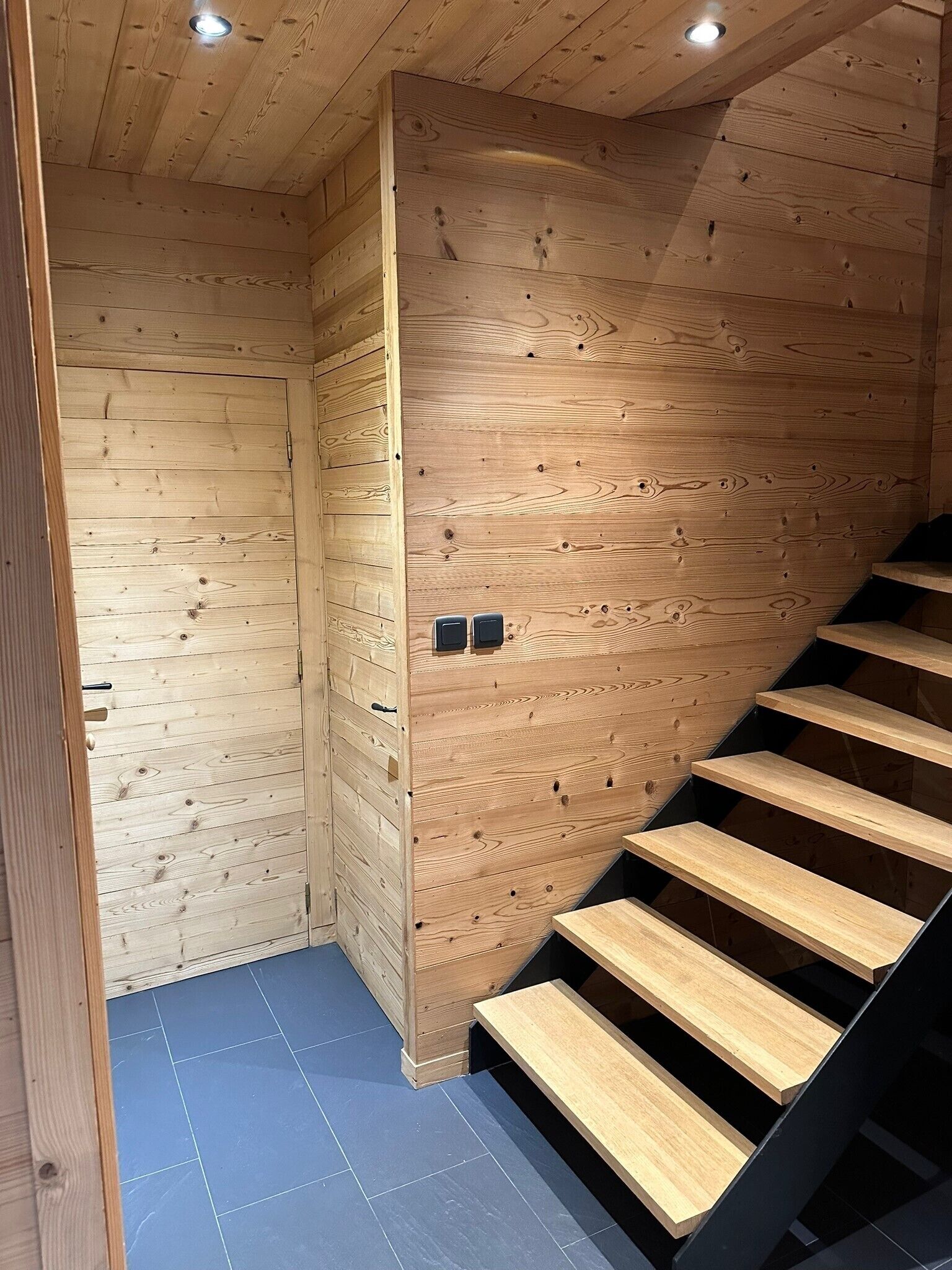 Un escalier en bois au design moderne, menant à une pièce dont un mur est en bois et dont le sol est recouvert de carreaux bleus.
