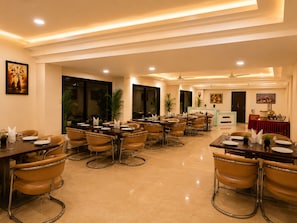 A hotel dining area with multiple tables and chairs, ceiling fans, and artwork on the walls. - TGI Jahaj Mahal Hotel  (Dhar)