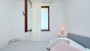 A bedroom with a bed, a nightstand, a lamp, a window with curtains, and a view of the sea. - Sunset Oasis (Dubrovnik)
