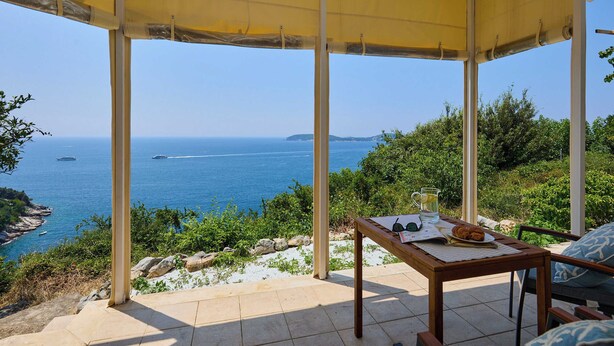 A covered outdoor seating area with a view of the sea, a table with a book, a glass of water, and a plate with food. - Sunset Oasis (Dubrovnik)
