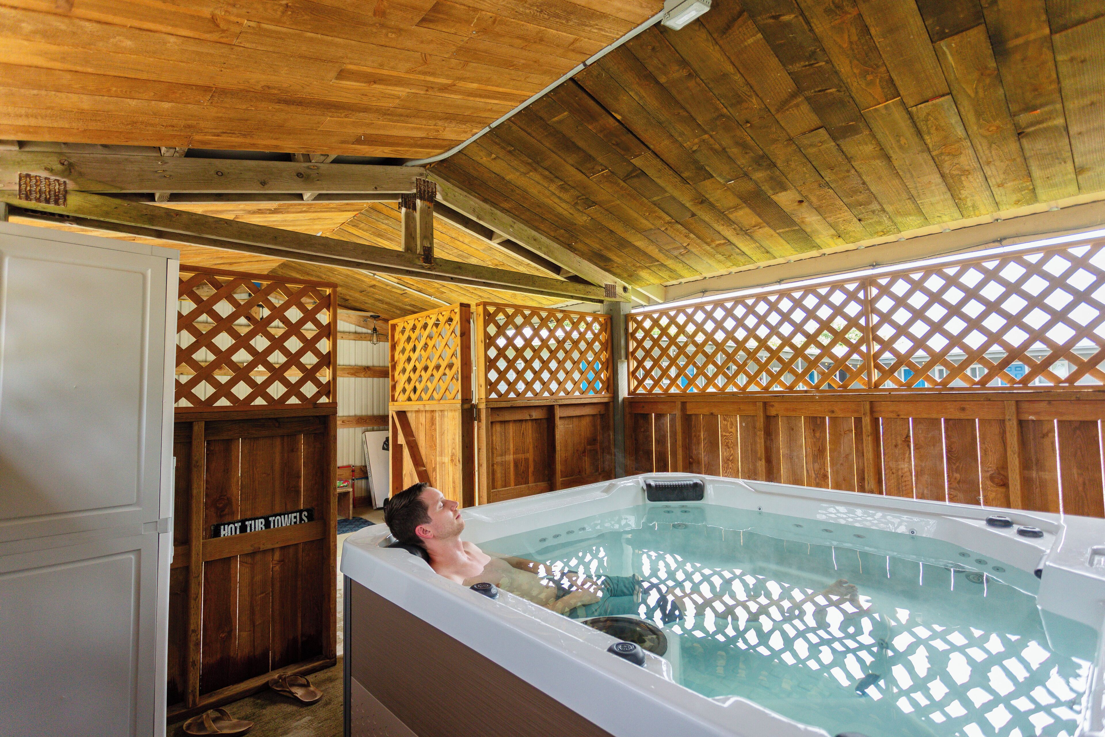 A man relaxing in a hot tub under a wooden ceiling with a wooden fence and a sign that reads "HOT TUBS".