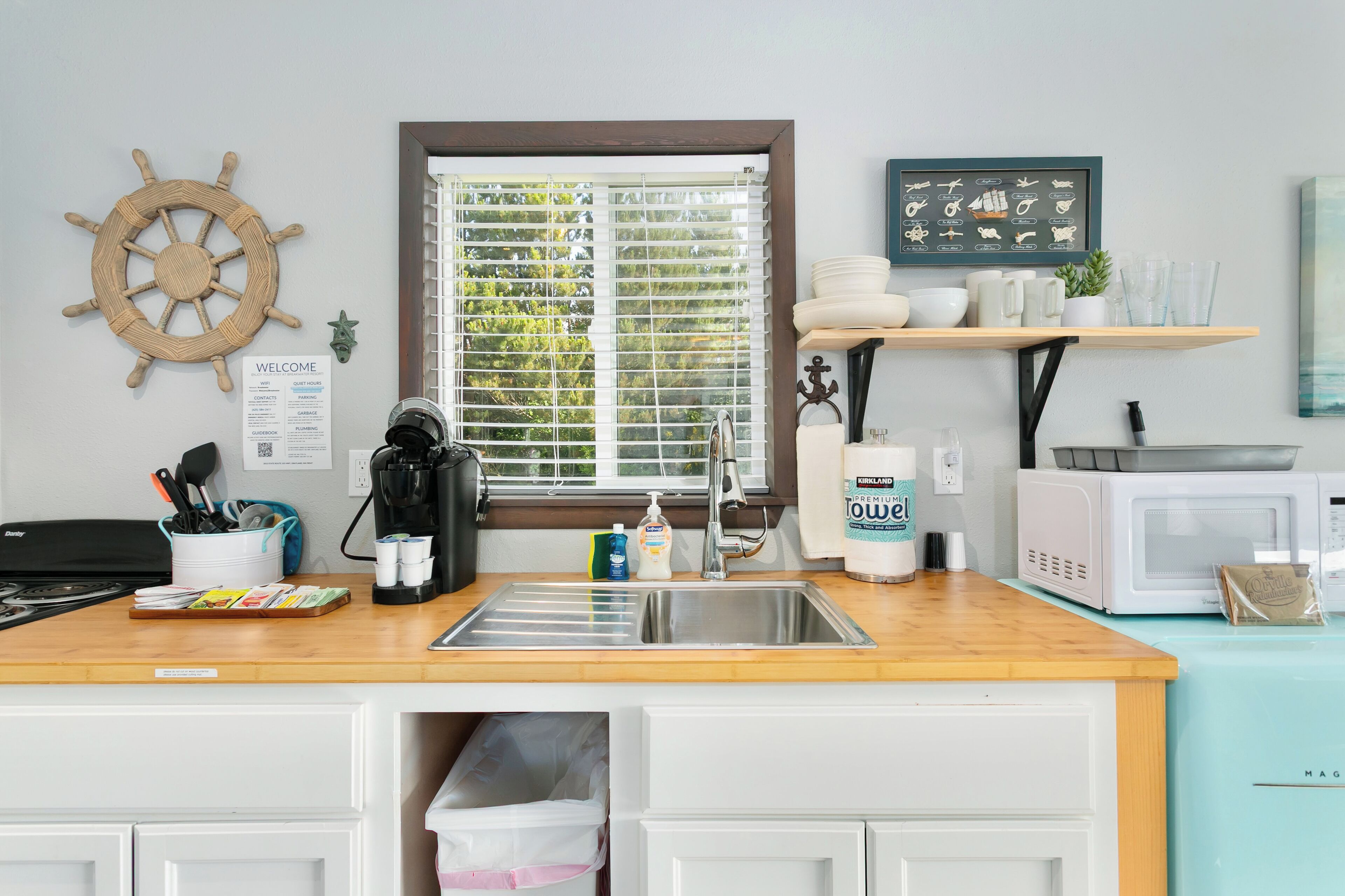 A kitchen with a wooden countertop, stainless steel sink, and white cabinets. There is a window with blinds, a ship's wheel decoration, and a framed nautical chart on the wall.