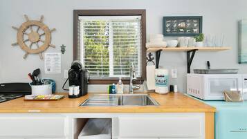 A kitchen with a wooden countertop, stainless steel sink, and white cabinets. There is a window with blinds, a ship's wheel decoration, and a framed nautical chart on the wall.