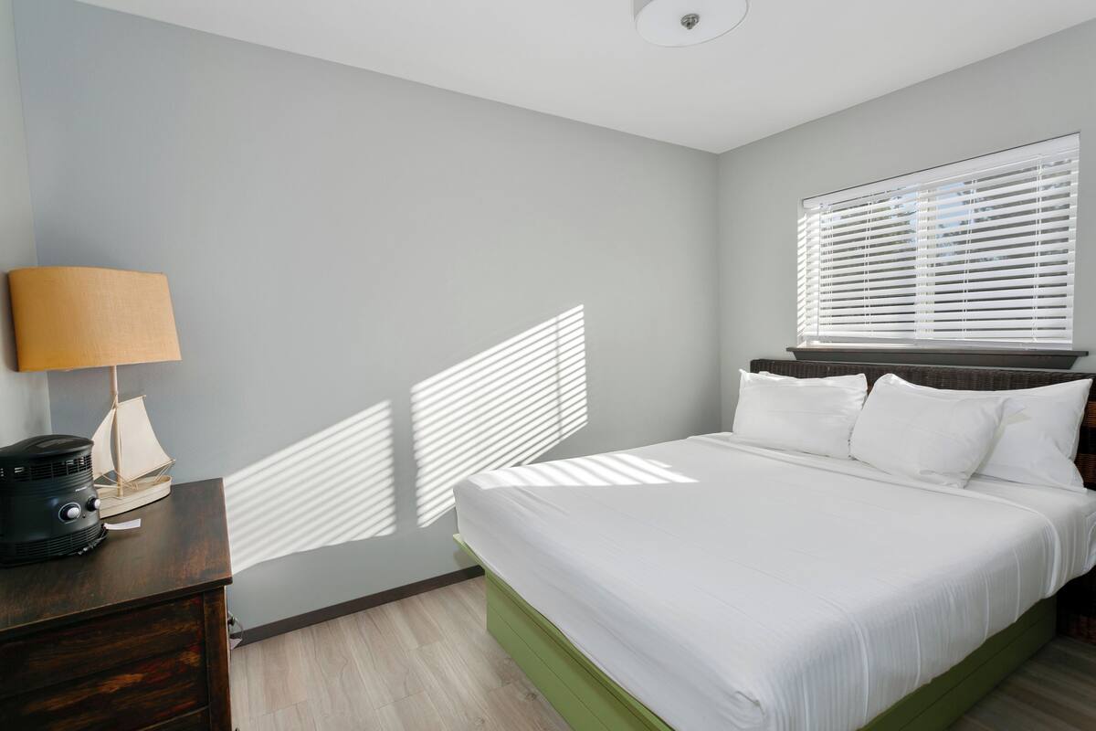 A bedroom with a bed, a dresser, a lamp, and a window with blinds.
