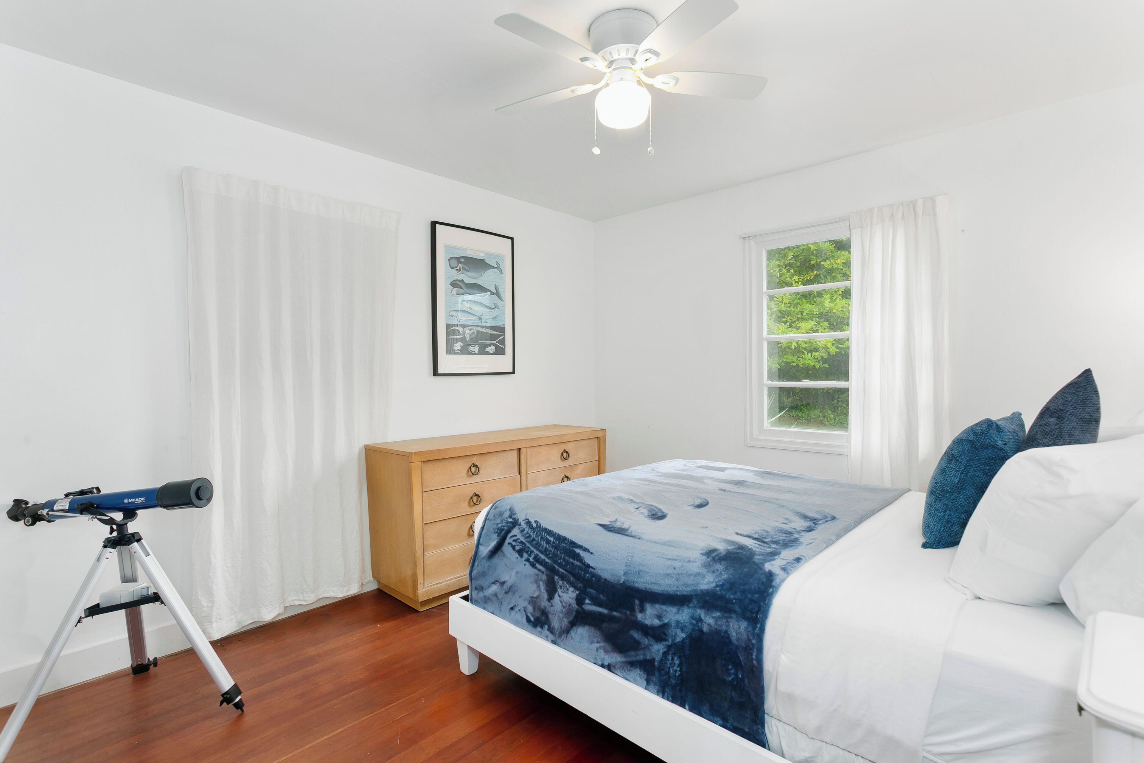 A bedroom with a bed, a wooden dresser, a telescope, and a window with curtains.
