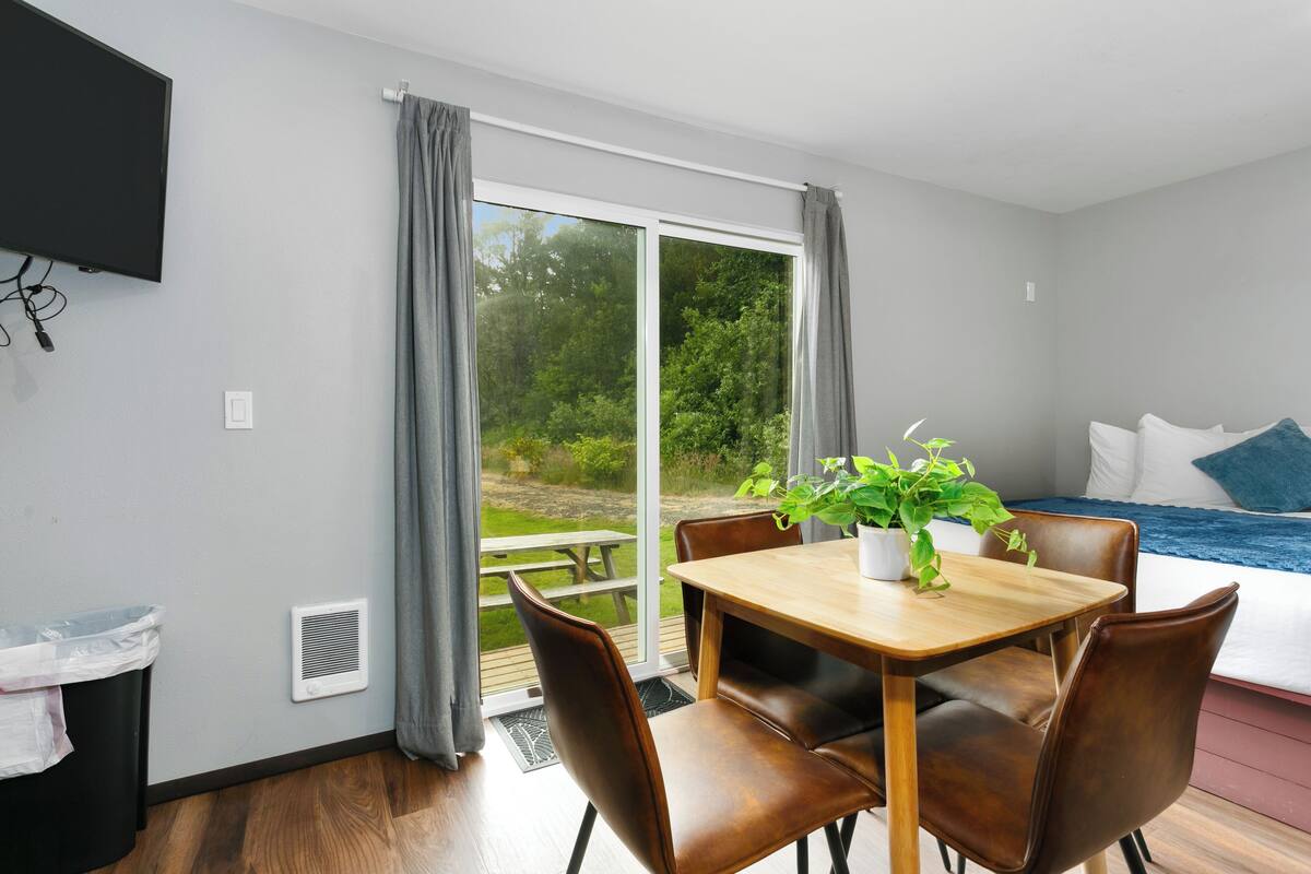 A modern room with a wooden dining table, leather chairs, a potted plant, a bed with a blue and white duvet, and a large window with curtains.