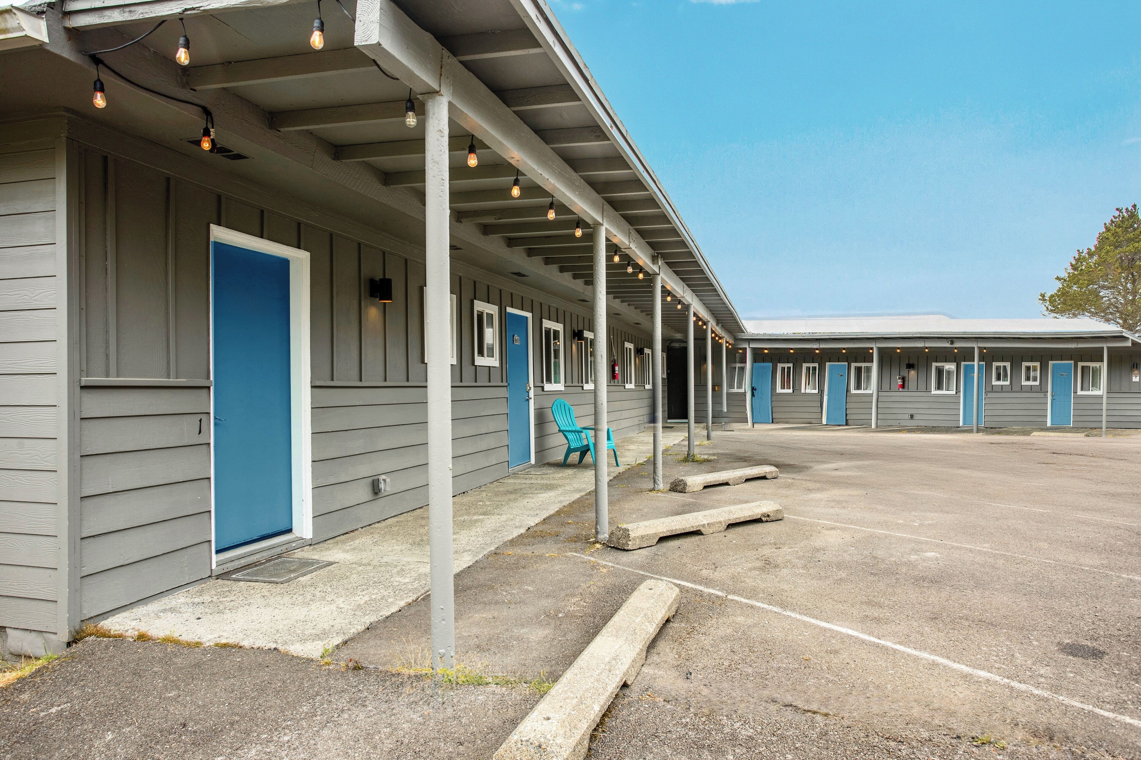 Un motel avec une rangée de chambres, chacune dotée d’une porte bleue et d’une fenêtre. Il y a un espace extérieur abrité avec un sol en béton et une chaise bleue.