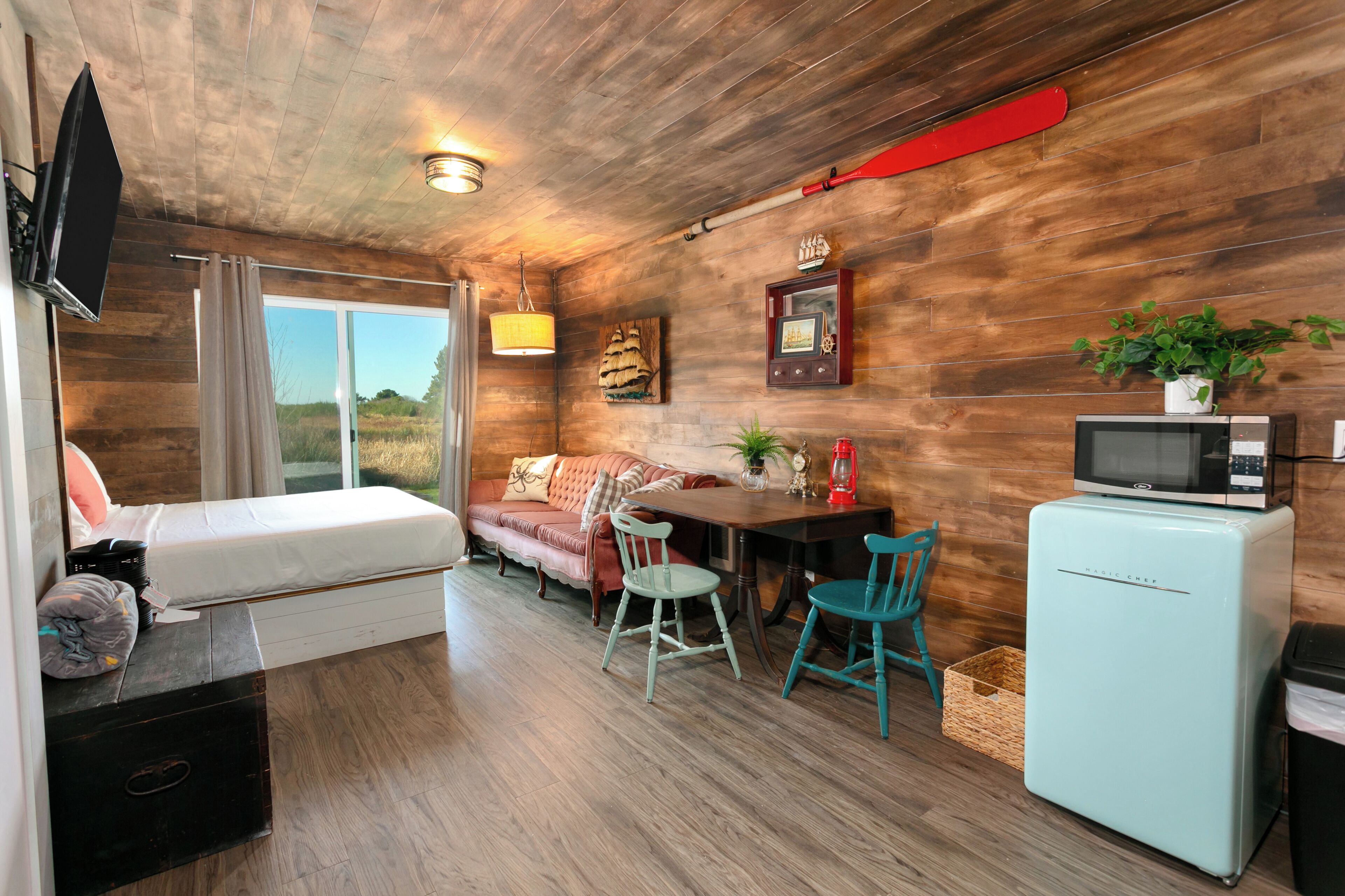 A compact room with a bed, a sofa, a dining table with chairs, a small fridge, and a TV mounted on the wall.