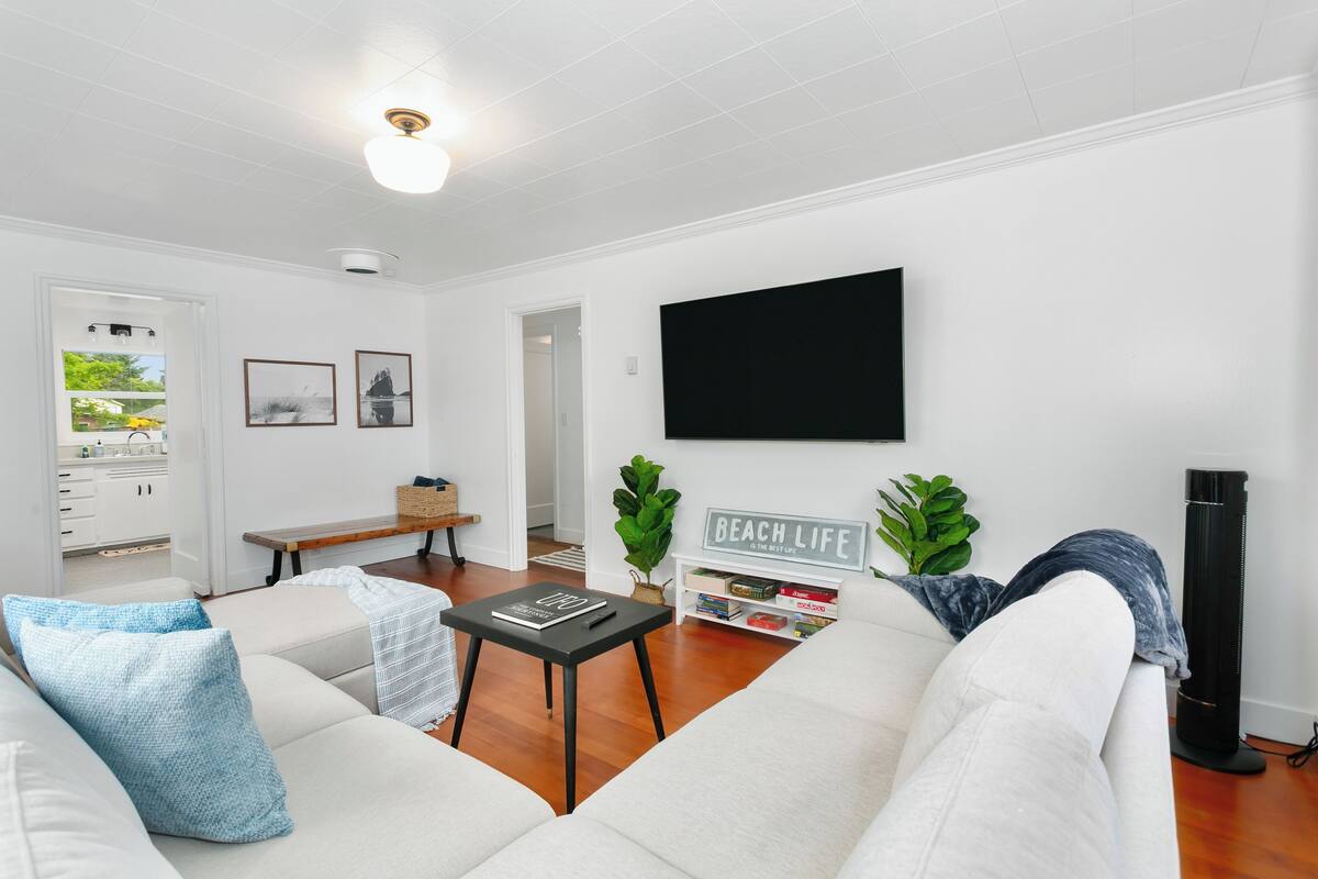 A living room with a white sofa, a black coffee table, and a flat-screen TV mounted on the wall. There is a bookshelf with books and a sign that says "BEACH LIFE".