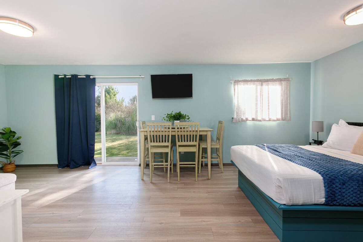 A bedroom with a bed, a desk with chairs, a TV, and a window with curtains.
