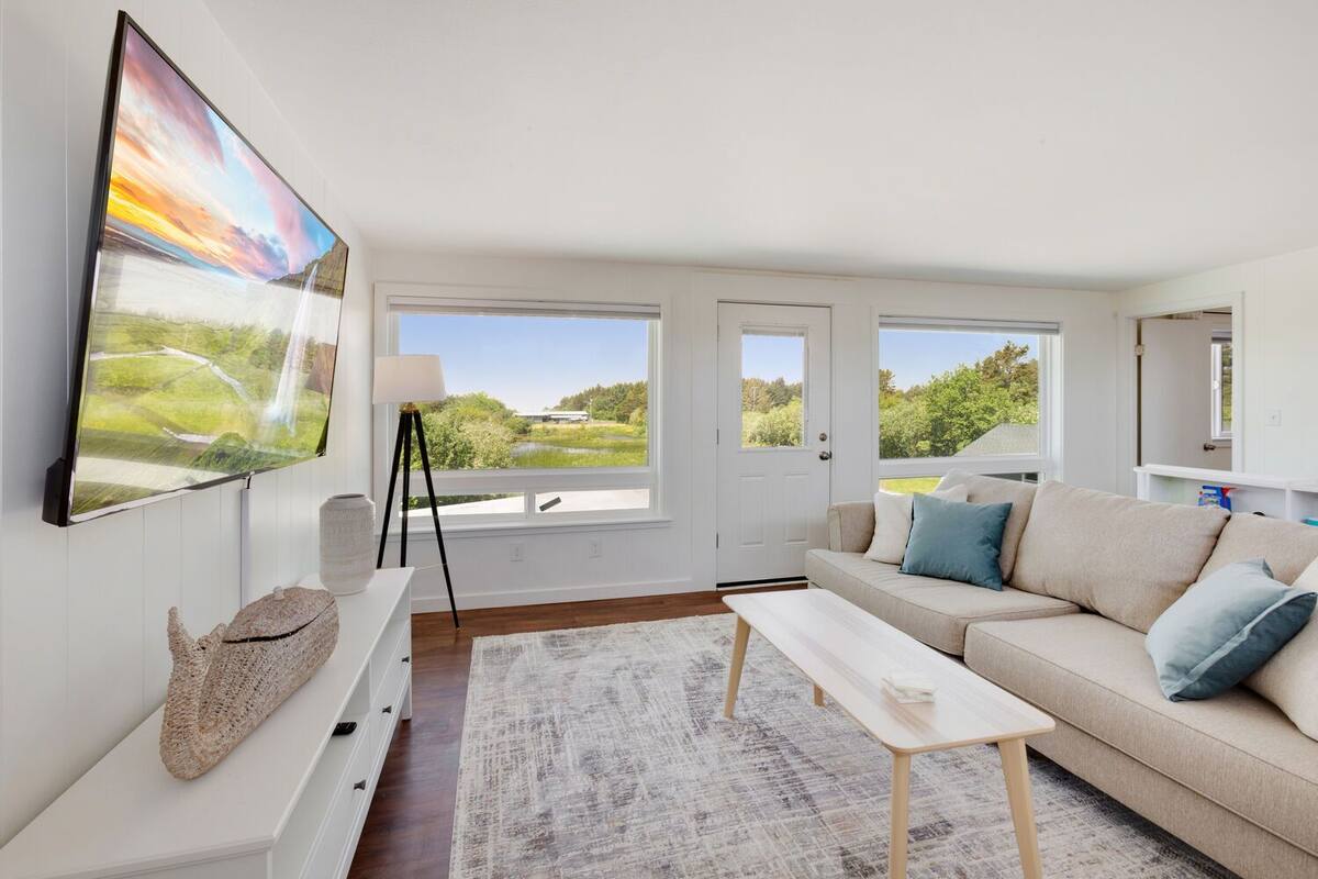 A modern living room with a large flat-screen TV, a sofa, a coffee table, and a view of a natural landscape through the windows.