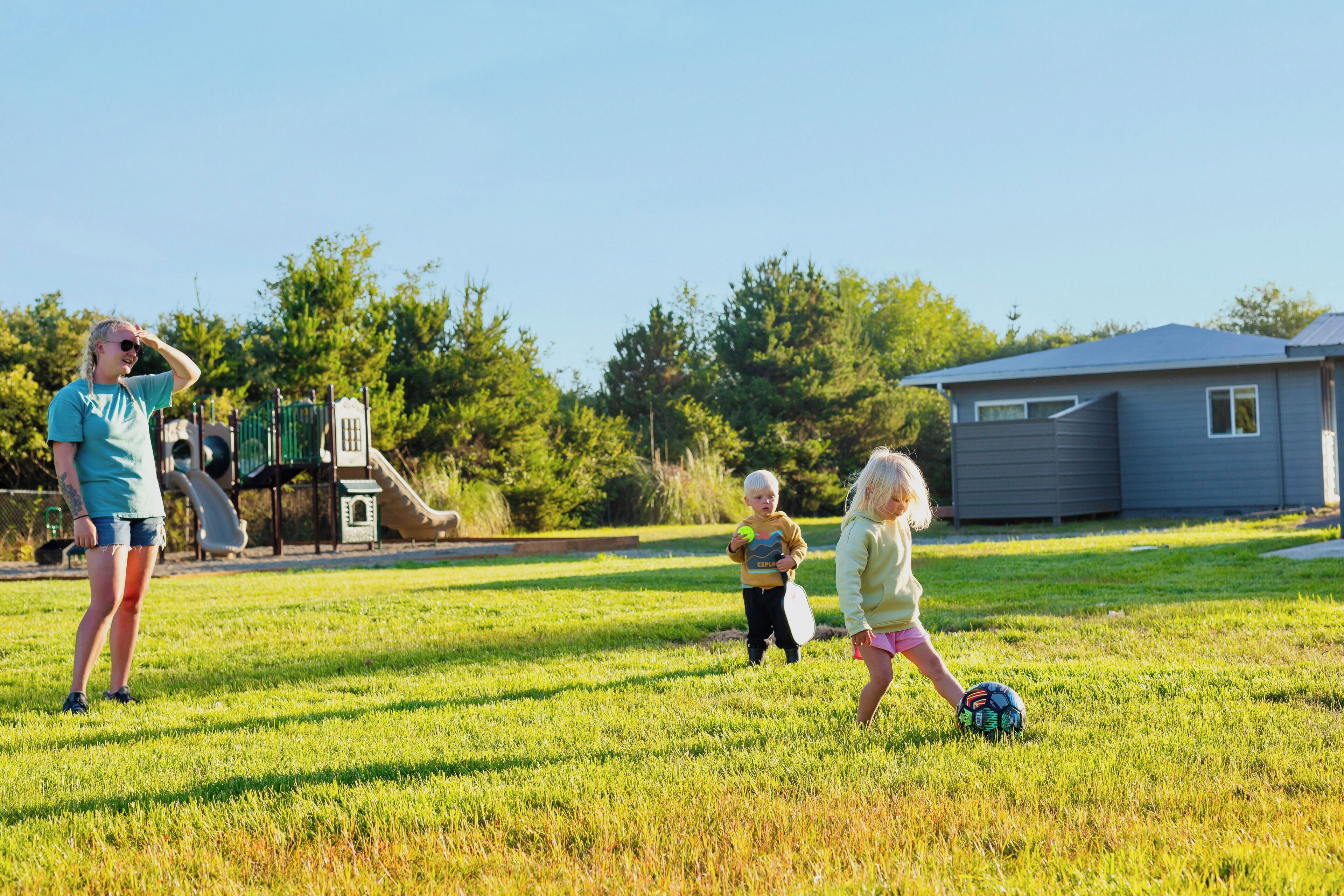 A grassy outdoor area with a playground structure, a house, and trees in the background.