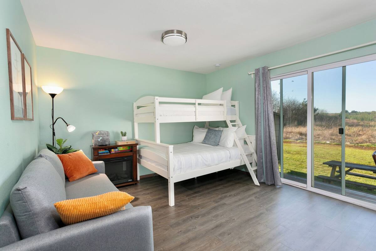 A room with a bunk bed, a sofa, a desk with a lamp, and a large sliding glass door to a garden.