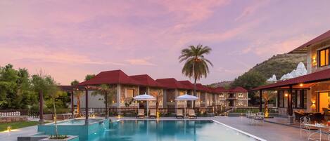 A swimming pool with lounge chairs and umbrellas, surrounded by buildings with red roofs and a mountainous backdrop.
