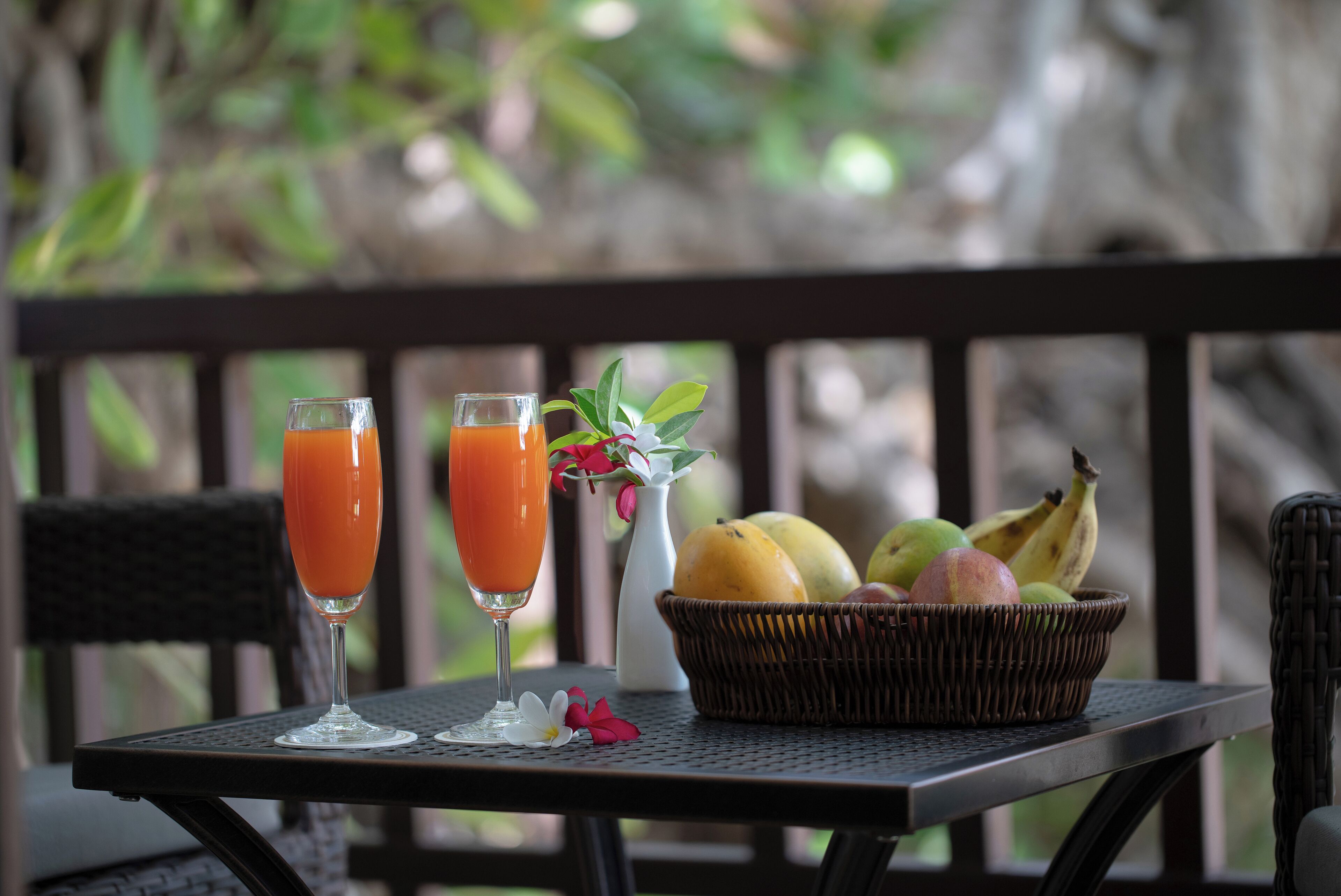 A table with two glasses of orange juice, a wicker basket with fruits, and a vase with flowers on a balcony.