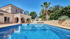 Ein Swimmingpool mit klarem, blauem Wasser, ein Haus mit einem Ziegeldach und im Hintergrund Palmen.