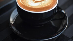 A cup of coffee with latte art on a black saucer on a reflective surface. - Roomshala 061 NK Residency (New Delhi)