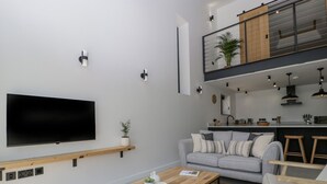 Un salon moderne doté d’un téléviseur à écran plat, d’une table basse en bois, d’un canapé gris et d’un espace cuisine situé à l’étage supérieur.