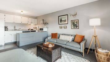 Een moderne keuken met witte kasten, een grijze bank met oranje kussens, een donkerhouten salontafel en een vloerlamp.