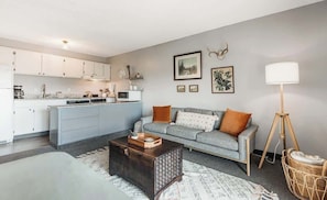 A modern kitchen with white cabinets, a grey sofa with orange cushions, a dark wooden coffee table, and a floor lamp. - Riverview Pet Friendly Condo in Downtown Bend (Bend)