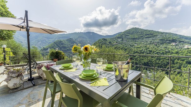 An outdoor dining area with a table set for four, surrounded by green chairs, a metal table lamp, and a large umbrella. - Belvedere – Avegno (Avegno)