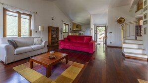 A spacious living room with a white sofa, a red sofa, and a wooden coffee table. There is a staircase with a golden railing leading to an upper level. - Belvedere – Avegno (Avegno)