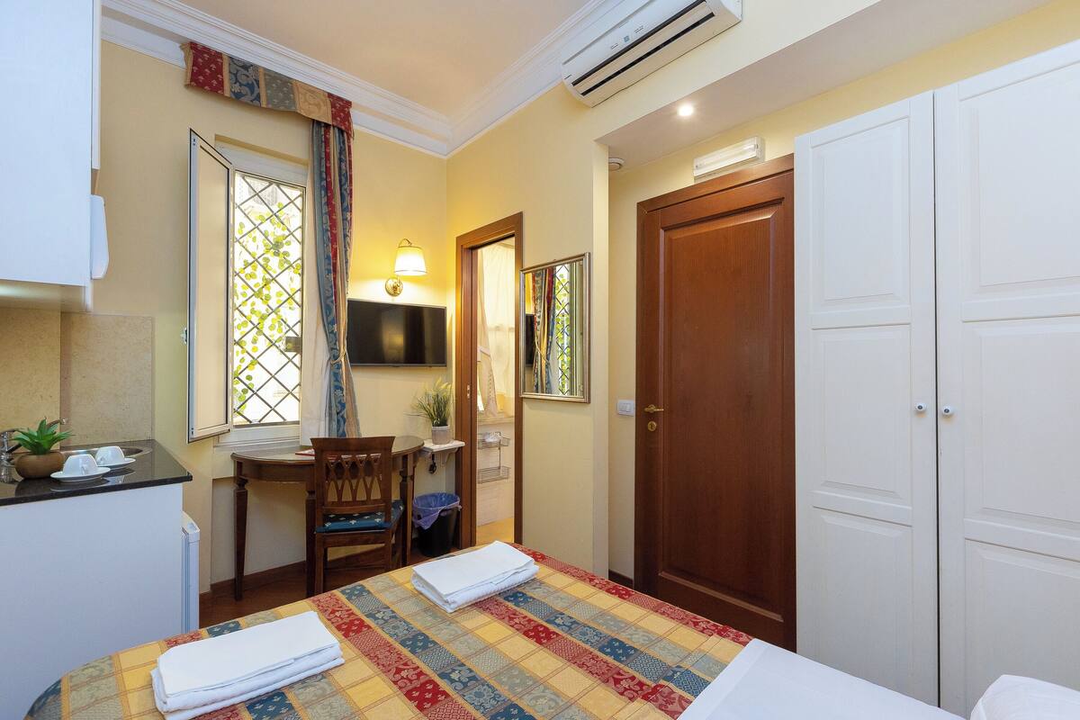 Quarto de hotel com uma cama, uma escrivaninha com cadeira, uma mesinha com planta, um espelho e um guarda-roupa.