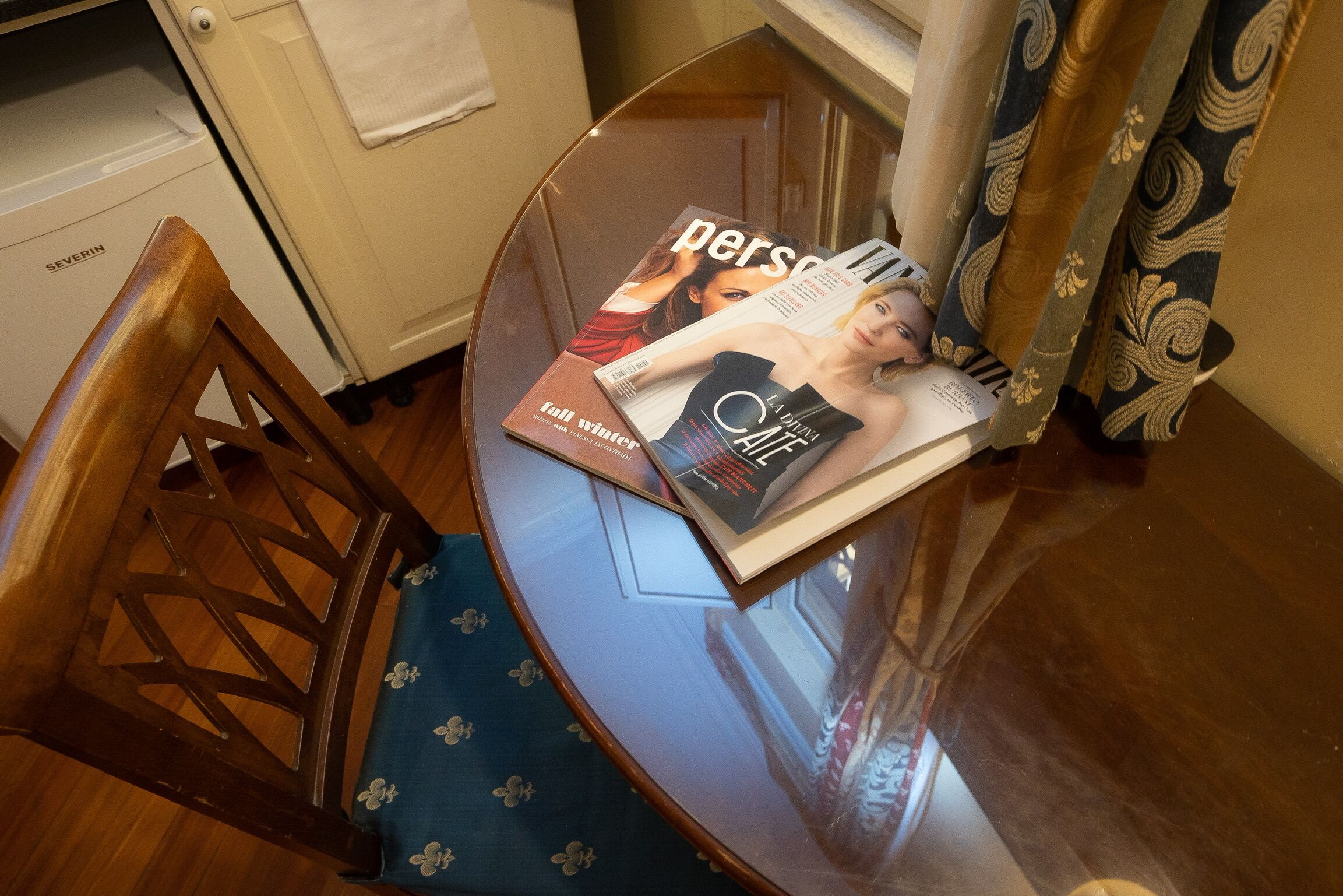 A magazine on a glass table with a chair beside it.