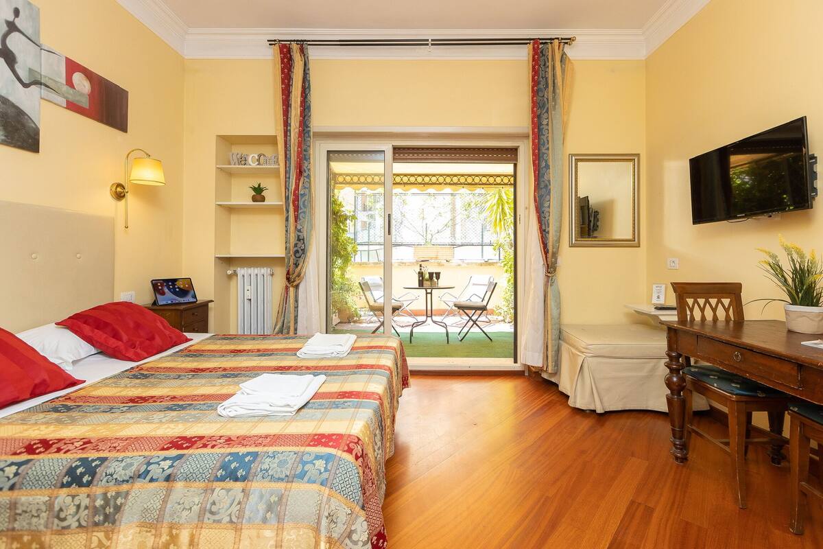Quarto de hotel com cama, escrivaninha, televisão e varanda com mesa e cadeiras.