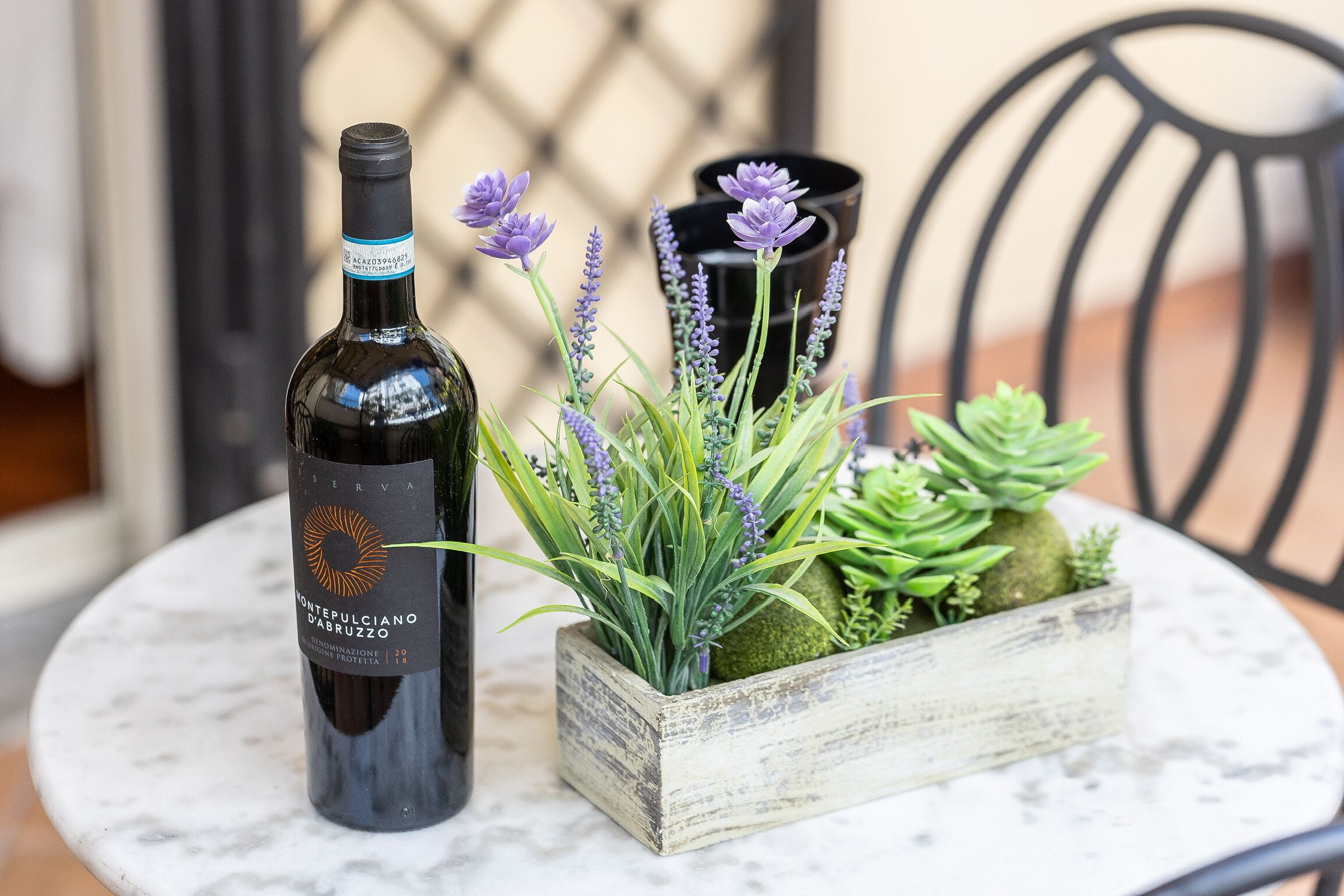 A bottle of wine, a wooden planter with lavender and succulents, and a black vase on a round table.