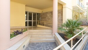 Ein Gebäudeeingang mit einer Treppe, die zu einer Glastür führt, einem kleinen Garten mit Pflanzen und einem Kübelpalmenbaum.