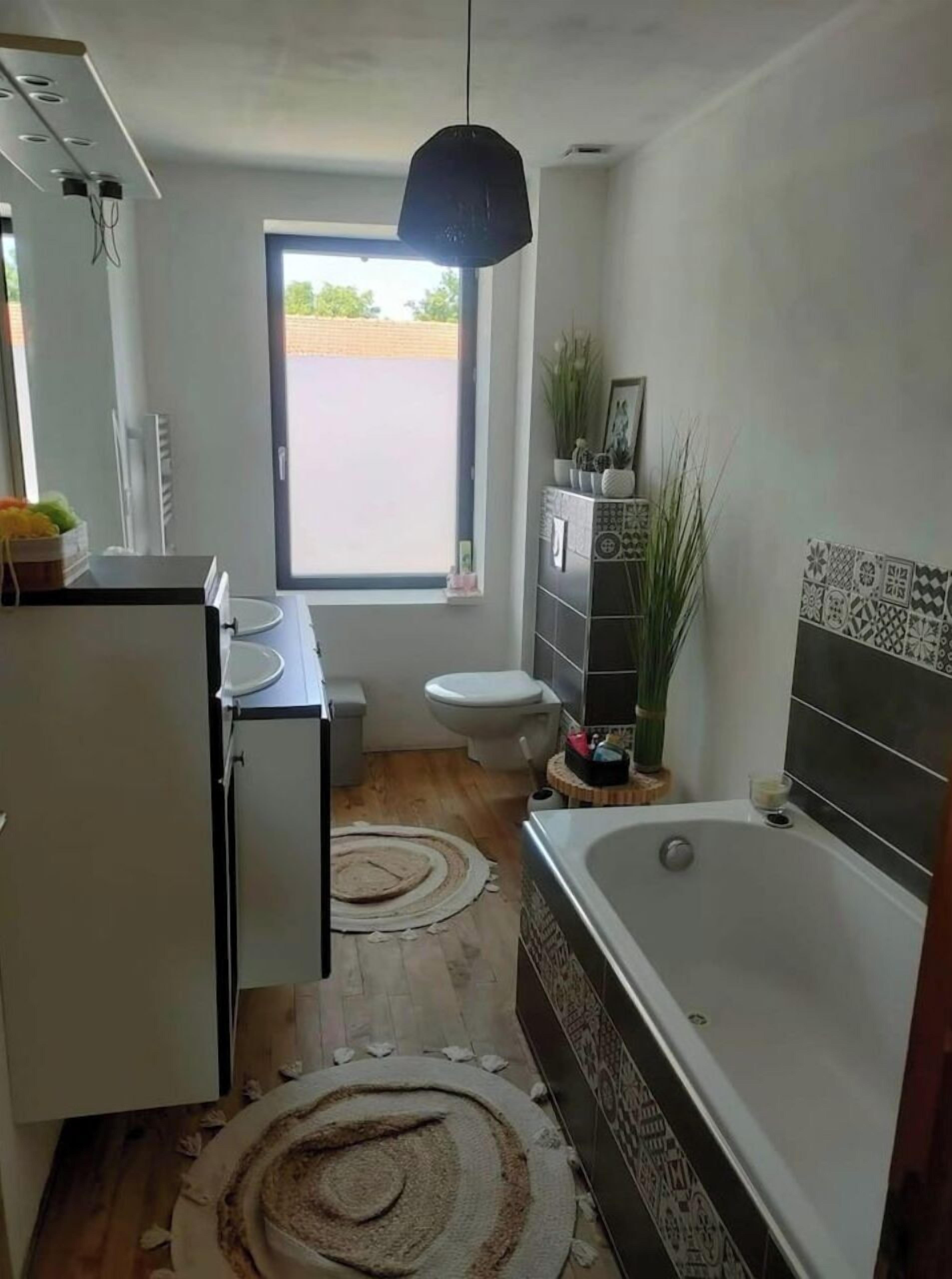 Une salle de bain moderne équipée d’une baignoire, de toilettes et d’un lavabo. Une fenêtre laisse entrer la lumière naturelle et offre une vue sur l’extérieur.