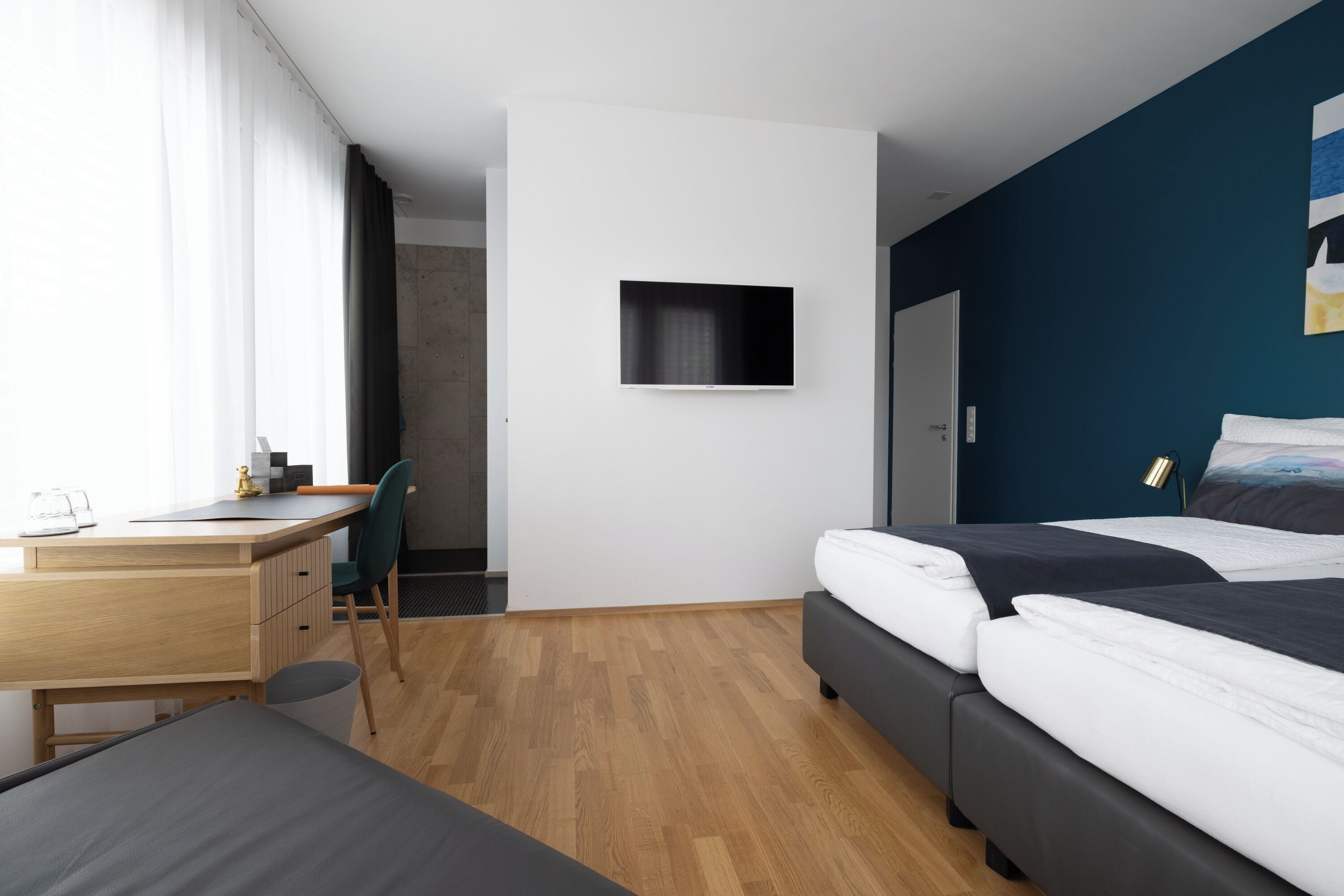 Una habitación de hotel moderna con un escritorio de madera, una cama con ropa oscura, un televisor montado y vistas de la ciudad a través de ventanas amplias.