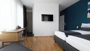 Una habitación de hotel moderna con un escritorio de madera, una cama con ropa oscura, un televisor montado y vistas de la ciudad a través de ventanas amplias.