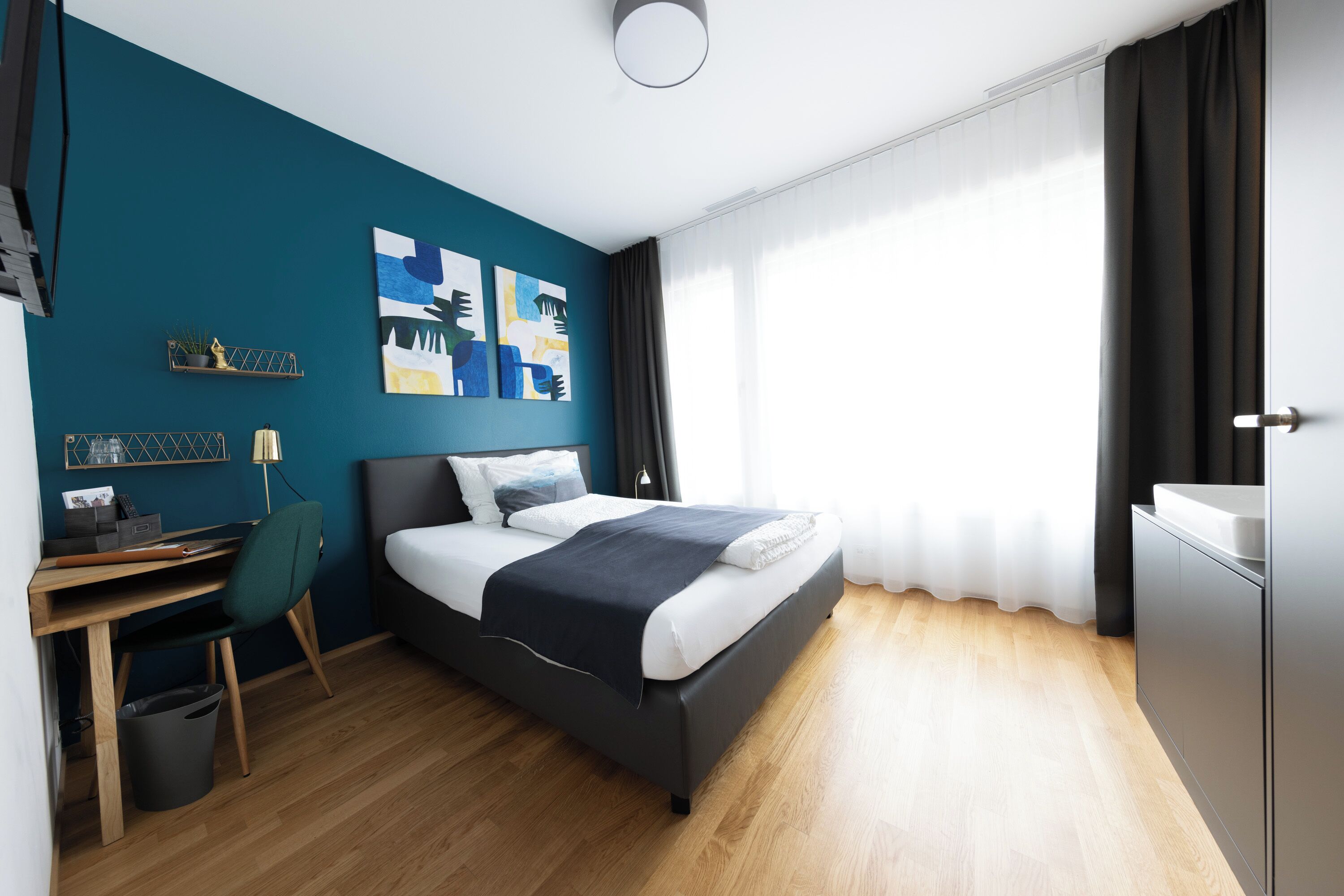 A modern hotel room with a bed, desk, and chair, a small trash bin, and a window with curtains.