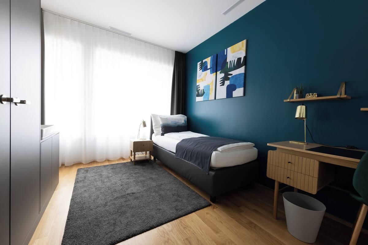 A modern bedroom with a bed, bedside table, desk, and a large window with curtains.