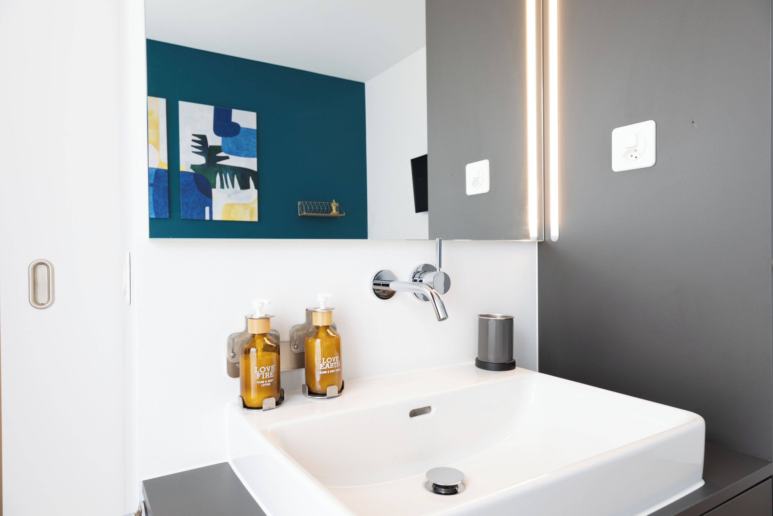 A modern bathroom with a white sink, two bottles of 'Love Earth' soap, a silver faucet, and a teal abstract painting on the wall.
