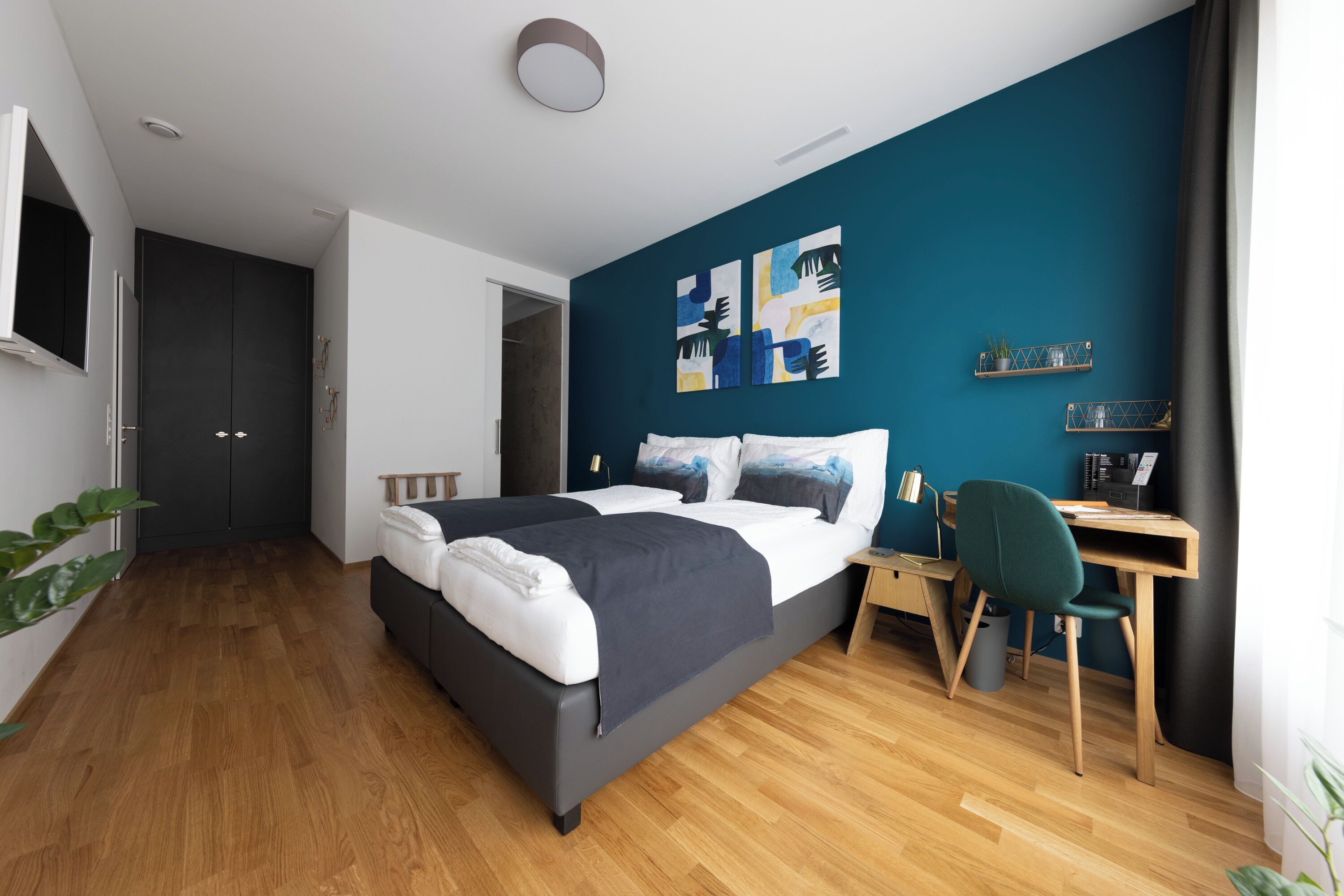 A modern bedroom with a bed, desk, and chair, featuring wooden flooring and a teal accent wall.