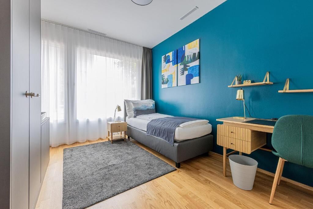 A bedroom with a bed, bedside table, lamp, desk, chair, and a blue accent wall.